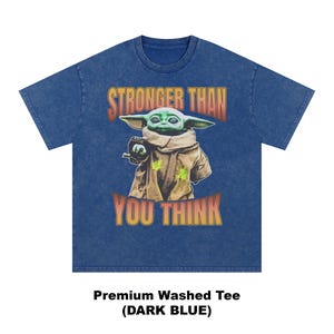 Pode incluir: Camiseta azul escuro com um gr&aacute;fico de Grogu, tamb&eacute;m conhecido como Baby Yoda, de The Mandalorian. A imagem inclui o texto "STRONGER THAN YOU THINK" em laranja e amarelo. A camisa &eacute; uma camiseta premium lavada.
