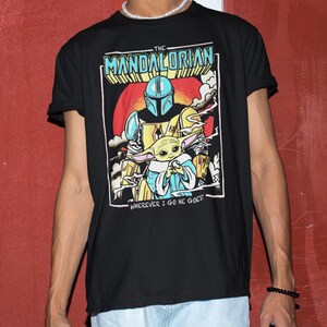 May include: Black t-shirt featuring a colorful graphic of The Mandalorian and Grogu. The design includes the text "THE MANDALORIAN" and "WHEREVER I GO HE GOES!" The artwork depicts the Mandalorian in armor holding Grogu, set against a red and orange background.