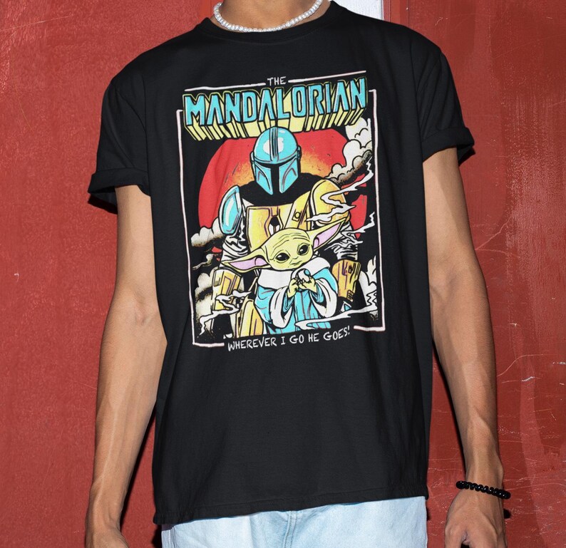 May include: Black t-shirt featuring a colorful graphic of The Mandalorian and Grogu. The design includes the text "THE MANDALORIAN" and "WHEREVER I GO HE GOES!" The artwork depicts the Mandalorian in armor holding Grogu, set against a red and orange background.