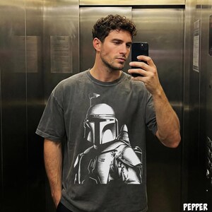 K&ouml;nnte beinhalten: Dunkelgraues T-Shirt mit einer detaillierten wei&szlig;en Grafik eines Mandalorianer-Helms und -R&uuml;stung. Das Shirt hat einen Vintage-Look und eine lockere Passform. Das Bild ist ein Selfie in einem Aufzug.