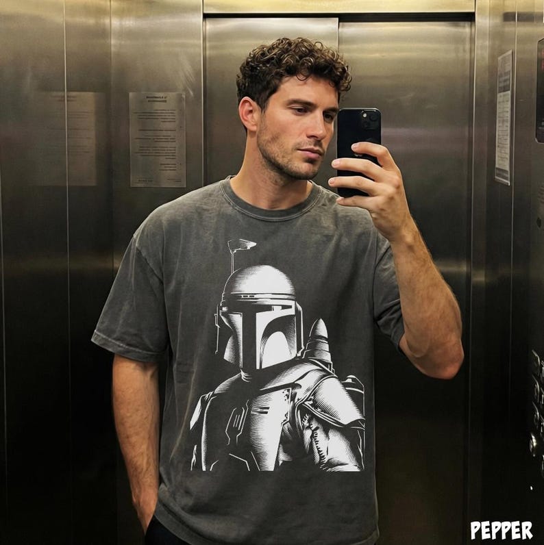 K&ouml;nnte beinhalten: Dunkelgraues T-Shirt mit einer detaillierten wei&szlig;en Grafik eines Mandalorianer-Helms und -R&uuml;stung. Das Shirt hat einen Vintage-Look und eine lockere Passform. Das Bild ist ein Selfie in einem Aufzug.