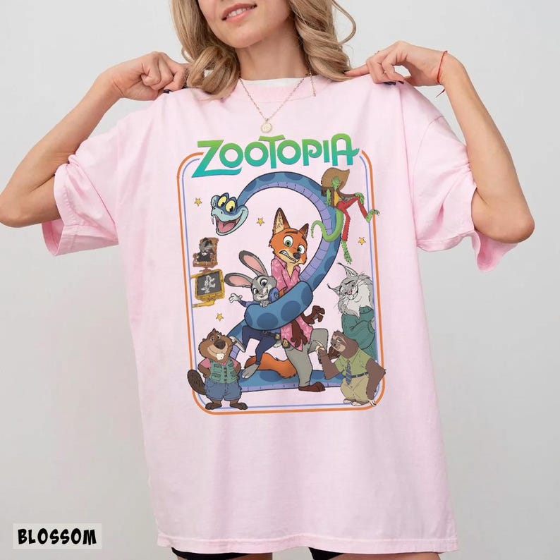 以下が含まれることがあります： 薄いピンク色のTシャツ。緑とオレンジで「ZOOTOPIA」の文字が描かれています。キツネ、ウサギ、ヘビなど、映画の漫画のキャラクターがデザインされています。「BLOSSOM」の文字が左下にプリントされています。