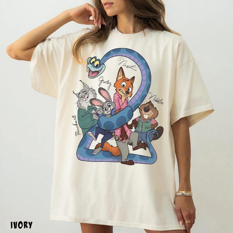 May include: Ivory-colored t-shirt featuring a cartoon graphic of characters from Zootopia. The design includes a blue snake with the names "Judy", "Nick", "Pawbert", and "Nibbles". The shirt has short sleeves and a crew neck.