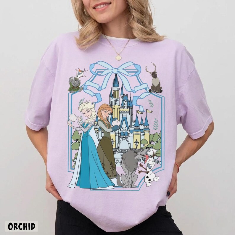 May include: Orchid-colored t-shirt featuring a graphic of Elsa and Anna from Frozen, with a castle backdrop and various characters. The design is framed by a blue ribbon. The word "ORCHID" is printed at the bottom.