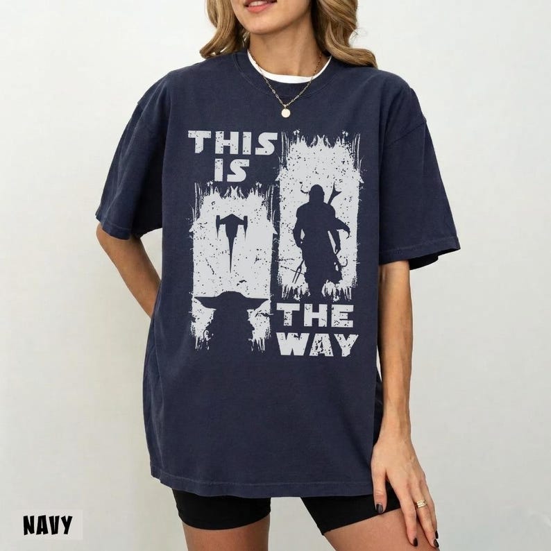 May include: Navy blue t-shirt with a distressed white graphic. The graphic features the text "THIS IS THE WAY" with images of a spaceship, a character, and a creature. The shirt is a relaxed fit.