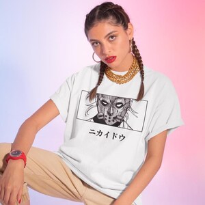 May include: White t-shirt featuring a black and white graphic of a masked character within a rectangular frame. Japanese text is below the graphic. The shirt has rolled-up sleeves.