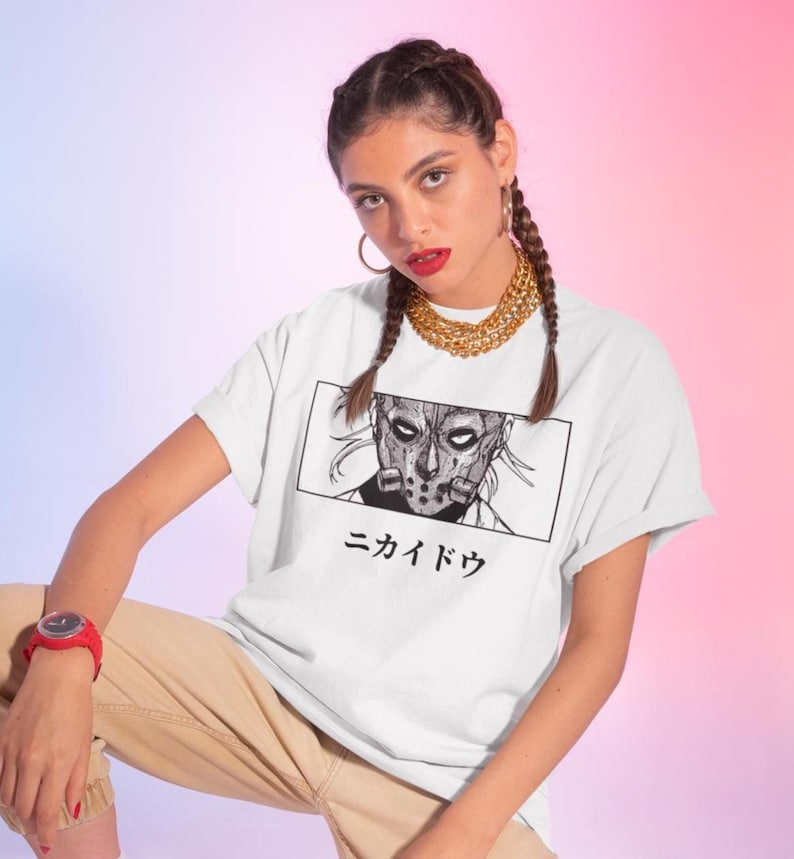 May include: White t-shirt featuring a black and white graphic of a masked character within a rectangular frame. Japanese text is below the graphic. The shirt has rolled-up sleeves.
