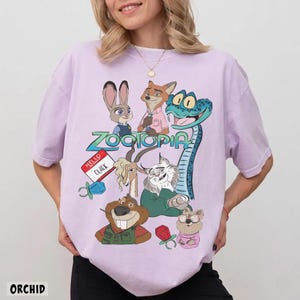 Może przedstawiać: Fioletowa koszulka z kolorową grafiką postaci z filmu animowanego Zwierzogr&oacute;d. Projekt zawiera Judy Hopps, Nicka Wilde i inne postacie. Słowo "Zwierzogr&oacute;d" jest wyświetlane w stylizowanej czcionce. Koszulka ma luźny kr&oacute;j.
