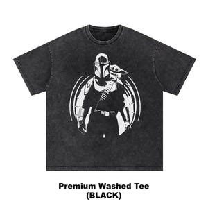 May include: Black washed tee with a white graphic of a Mandalorian character and Grogu, inside a circular design. The text "Premium Washed Tee (BLACK)" is printed at the bottom. The shirt has a crew neck and short sleeves.
