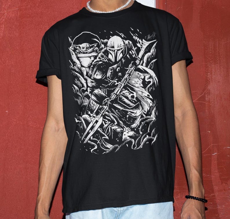 Puede incluir: Camiseta negra con un dise&ntilde;o gr&aacute;fico blanco detallado de un guerrero Mandaloriano y Grogu. El dise&ntilde;o incluye el casco y el arma del Mandaloriano, y a Grogu en una c&aacute;psula. Las mangas de la camiseta est&aacute;n dobladas.