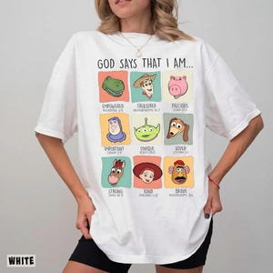 May include: White t-shirt with the text "GOD SAYS THAT I AM..." and colorful cartoon character illustrations. Each character is in a square with a descriptive word and a Bible verse reference.