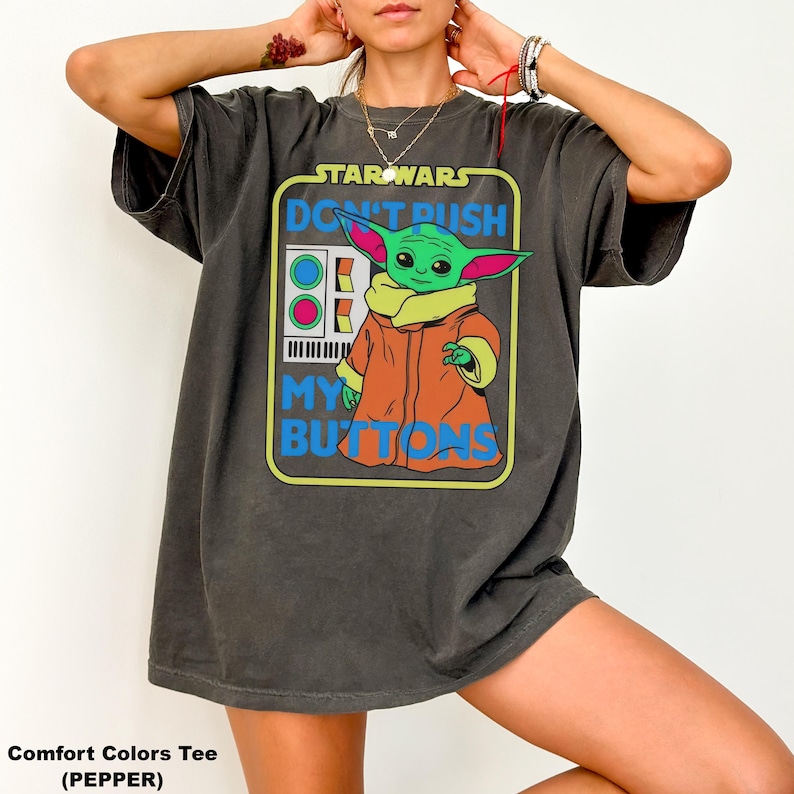 May include: Dark grey t-shirt featuring a graphic of Grogu from Star Wars with the text "DON'T PUSH MY BUTTONS". The graphic includes a green Grogu wearing an orange coat. The shirt is a Comfort Colors Tee in Pepper.