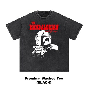 Op de afbeelding: Zwart premium gewassen T-shirt met een afbeelding van The Mandalorian en Grogu. Het ontwerp bevat de helm van de Mandalorian en Grogu in wit, met de tekst "THE MANDALORIAN and Grogu" in rood. De stijl van het shirt is een vintage wassing.