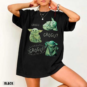 May include: Black t-shirt featuring four images of Grogu from The Mandalorian. The images are in green and white, with the words "GROGU...", "GROGU?", and "GROGU!" printed above and below the images. The word "BLACK" is printed at the bottom.