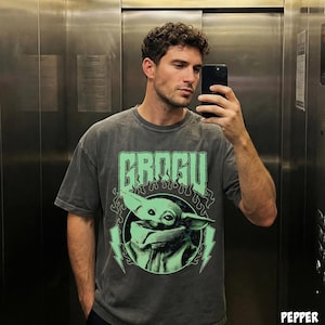 May include: Dark gray t-shirt featuring the word "GROGU" in a stylized font above a green image of Grogu, with lightning bolts and flames. The shirt is a short-sleeved, crew neck style.