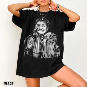 Puede incluir: Camiseta negra con un gr&aacute;fico en blanco y negro de un hombre y una peque&ntilde;a criatura alien&iacute;gena. El hombre lleva una armadura y el alien&iacute;gena est&aacute; envuelto en una capa. La palabra "BLACK" est&aacute; impresa en la parte inferior.
