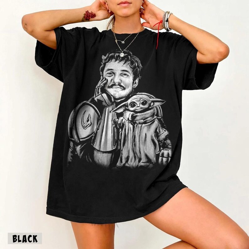 Puede incluir: Camiseta negra con un gr&aacute;fico en blanco y negro de un hombre y una peque&ntilde;a criatura alien&iacute;gena. El hombre lleva una armadura y el alien&iacute;gena est&aacute; envuelto en una capa. La palabra "BLACK" est&aacute; impresa en la parte inferior.