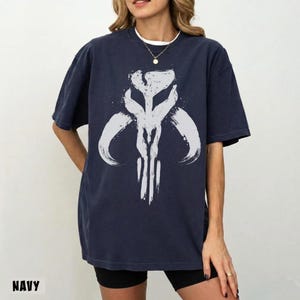 May include: Navy blue t-shirt featuring a white, stylized graphic design. The design appears to be a symbol or emblem, with brushstroke-like details. The word "NAVY" is printed at the bottom left corner of the shirt.