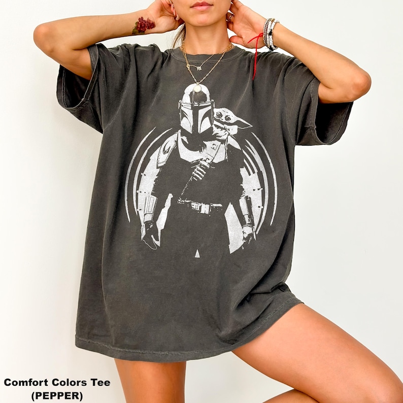 May include: Dark grey Comfort Colors tee featuring a white graphic of a Mandalorian character and Grogu. The design is set within a circular frame. The shirt has short sleeves and a relaxed fit. The text "Comfort Colors Tee (PEPPER)" is printed at the bottom.