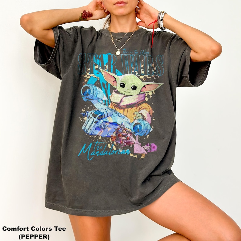 May include: Dark grey t-shirt with a graphic of Grogu from The Mandalorian, featuring the text "STAR WARS" and "The Mandalorian". The design includes a spaceship and Grogu in a yellow robe. The shirt is a Comfort Colors Tee.