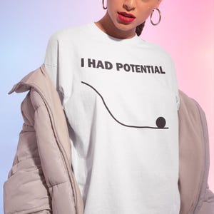 May include: White t-shirt with the text "I HAD POTENTIAL" in black, above a simple black line drawing of a ball rolling down a hill. The shirt is part of a clothing collection.
