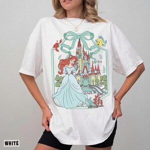 May include: White t-shirt featuring a colorful graphic of a princess in a blue dress, with a castle and sea creatures. The design is framed by a green border and bow. The word "WHITE" is printed at the bottom left.
