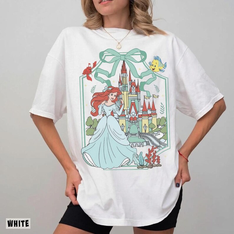 May include: White t-shirt featuring a colorful graphic of a princess in a blue dress, with a castle and sea creatures. The design is framed by a green border and bow. The word "WHITE" is printed at the bottom left.