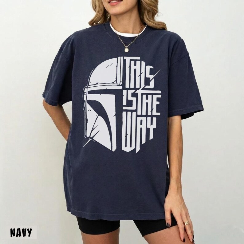 May include: Navy blue t-shirt with a white graphic of a helmet and the text "THIS IS THE WAY". The design is centered on the front of the shirt. The shirt has a relaxed fit.