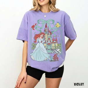 May include: A violet-colored t-shirt featuring a graphic of a princess in a blue gown, a castle, and sea creatures. The design is framed by a green bow and line art. The word "VIOLET" is printed in white at the bottom right.