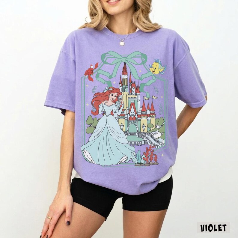 May include: A violet-colored t-shirt featuring a graphic of a princess in a blue gown, a castle, and sea creatures. The design is framed by a green bow and line art. The word "VIOLET" is printed in white at the bottom right.