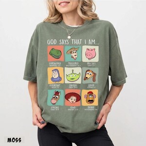 May include: A moss green t-shirt with the text "GOD SAYS THAT I AM..." and cartoon character illustrations. The shirt features various characters with words like "Empowered," "Treasured," and "Precious."