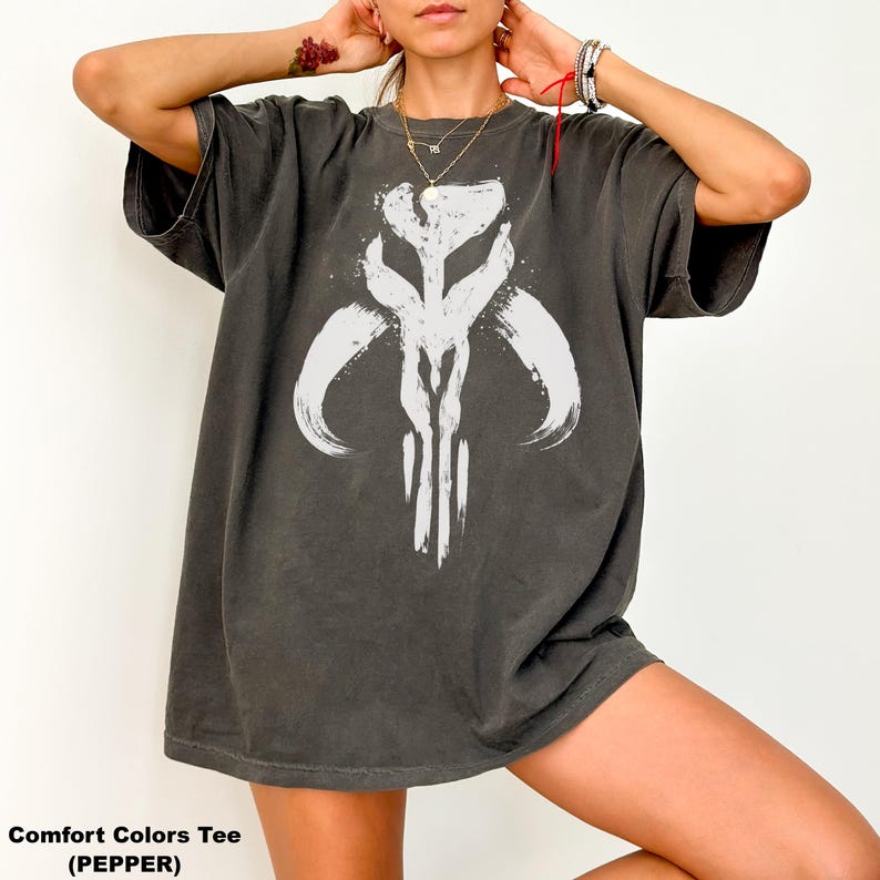 May include: Dark gray Comfort Colors tee shirt with a white graphic design. The design is a stylized, abstract symbol. The shirt is a relaxed fit and has short sleeves. The text "Comfort Colors Tee (PEPPER)" is at the bottom of the image.