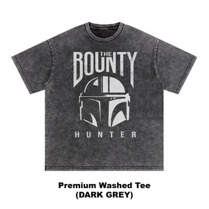 Puede incluir: Camiseta gris oscuro con un gr&aacute;fico desgastado. El dise&ntilde;o presenta las palabras "THE BOUNTY HUNTER" sobre un casco estilizado. La camiseta es una camiseta lavada de primera calidad.