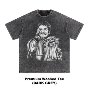 Puede incluir: Camiseta gris oscuro lavado con una ilustraci&oacute;n en blanco y negro de un hombre y una peque&ntilde;a criatura alien&iacute;gena. El hombre lleva una armadura y el alien&iacute;gena est&aacute; envuelto en una capa. El texto "Premium Washed Tee (DARK GREY)" est&aacute; impreso en la parte inferior.