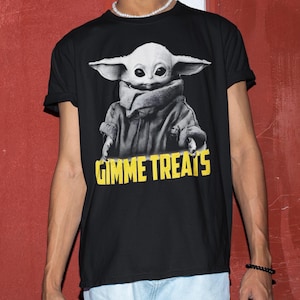 Op de afbeelding: Zwart T-shirt met een grijswaardenafbeelding van een personage met grote oren en de tekst "GIMME TREATS" in het geel. Het personage draagt een jas en een sjaal. Ronde hals.