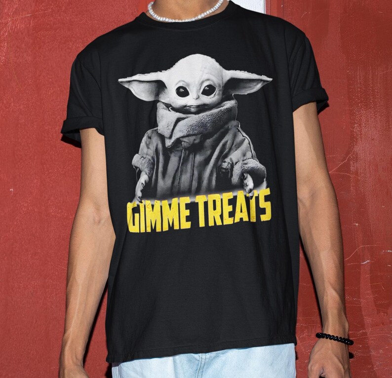 Op de afbeelding: Zwart T-shirt met een grijswaardenafbeelding van een personage met grote oren en de tekst "GIMME TREATS" in het geel. Het personage draagt een jas en een sjaal. Ronde hals.