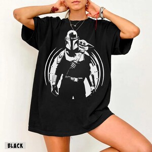 May include: Black t-shirt with a white graphic of The Mandalorian and Grogu within a white circle. The word "BLACK" is printed at the bottom left. The shirt has short sleeves and a loose fit.