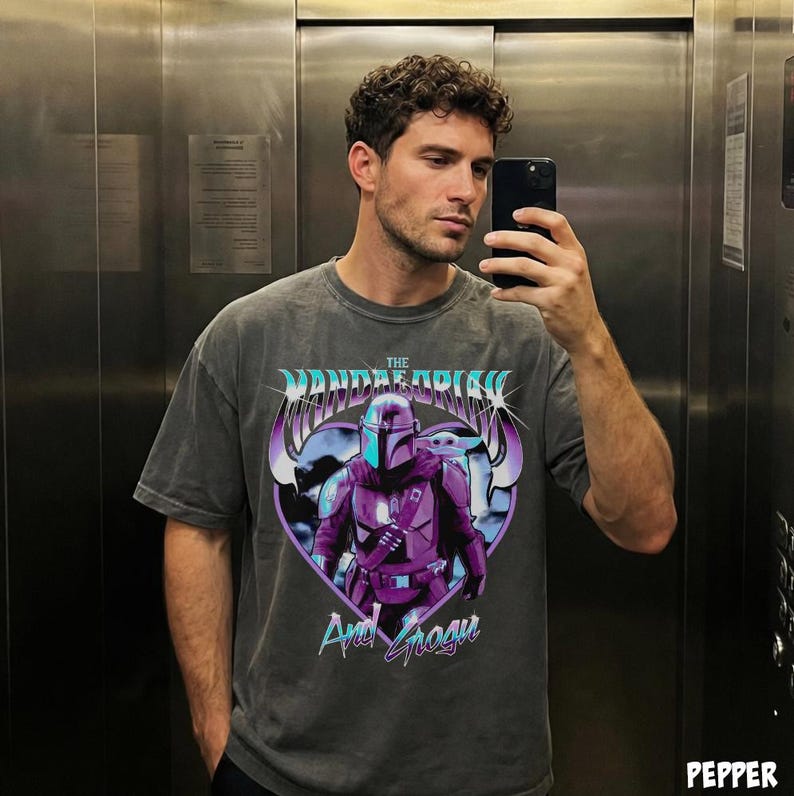 May include: A gray t-shirt featuring a graphic of The Mandalorian and Grogu in a heart-shaped design. The text "The Mandalorian" is arched above the image, with "And Grogu" below. The shirt has a vintage, worn look.