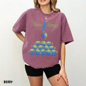 May include: Berry-colored t-shirt featuring the text "Pizza Planet" in green, with a graphic of the Little Green Men from Toy Story. The design includes a claw and a pyramid of aliens. The shirt is a comfortable, oversized fit.