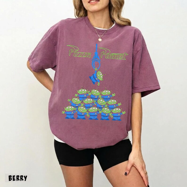 May include: Berry-colored t-shirt featuring the text "Pizza Planet" in green, with a graphic of the Little Green Men from Toy Story. The design includes a claw and a pyramid of aliens. The shirt is a comfortable, oversized fit.