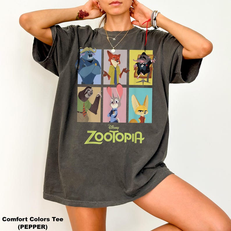 May include: Dark gray Comfort Colors tee featuring a colorful graphic of characters from the Disney movie Zootopia. The shirt has a crew neck and short sleeves. The word "Zootopia" is printed in green and white below the graphic.