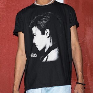 Op de afbeelding: Zwart T-shirt met een zwart-wit profielafbeelding. Het Star Wars-logo is links onderin in het wit gedrukt. Het shirt heeft korte mouwen.