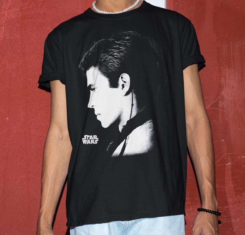 Op de afbeelding: Zwart T-shirt met een zwart-wit profielafbeelding. Het Star Wars-logo is links onderin in het wit gedrukt. Het shirt heeft korte mouwen.