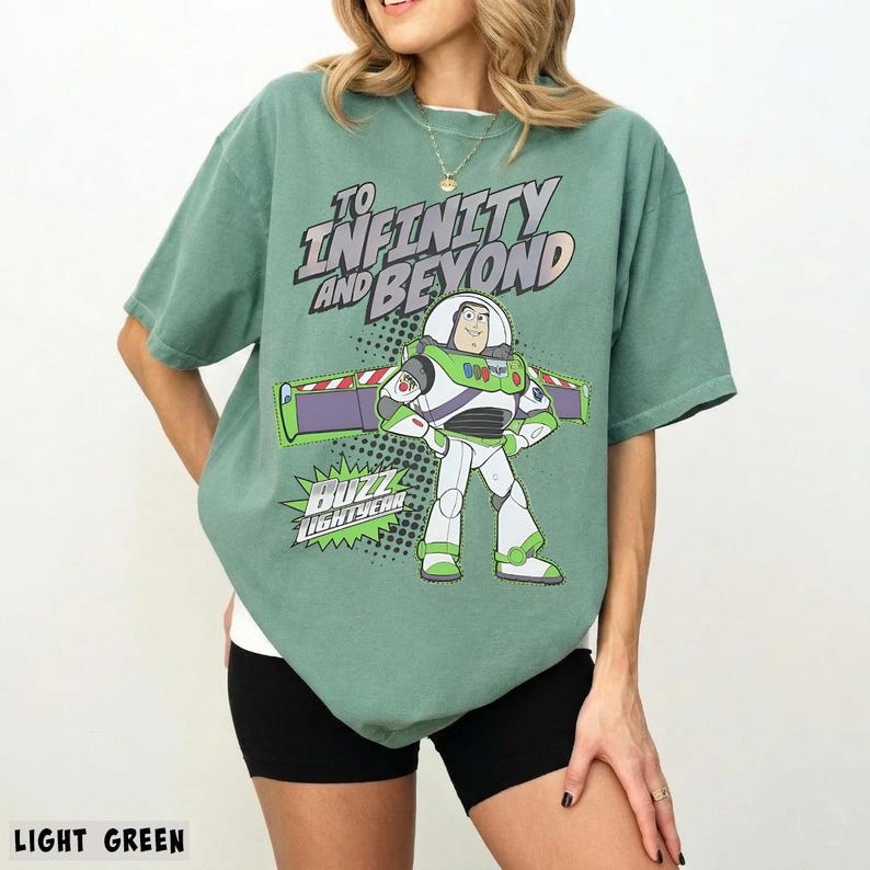 May include: A light green short-sleeve t-shirt featuring a graphic of the character Buzz Lightyear. The text reads TO INFINITY AND BEYOND above the character and BUZZ LIGHTYEAR below. The design includes a purple and green wing element.