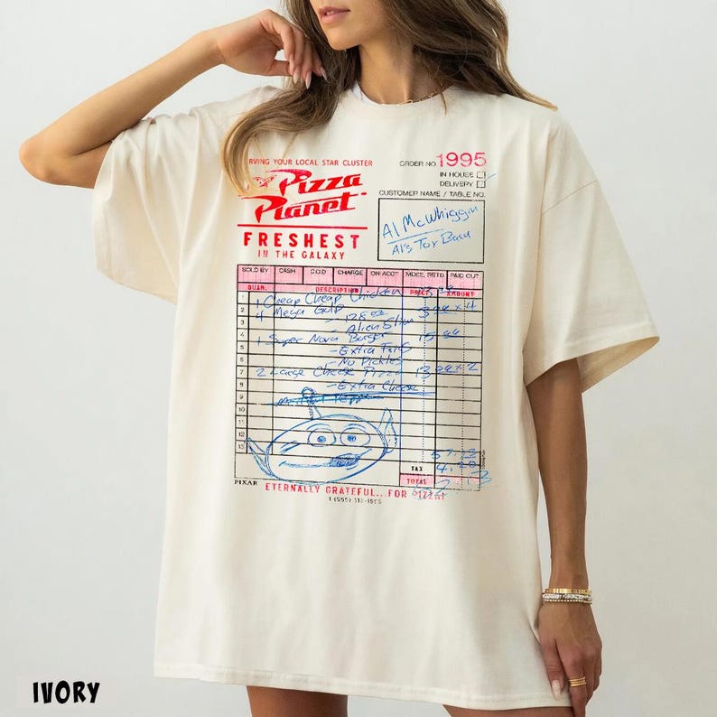 Puede incluir: Camiseta de color marfil con el logotipo de Pizza Planet y un gr&aacute;fico estilo recibo. El gr&aacute;fico incluye el texto "Freshest in the Galaxy" y un personaje alien&iacute;gena, con notas manuscritas.