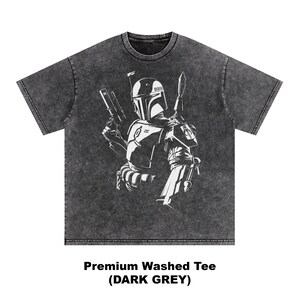 May include: Dark grey washed tee featuring a white graphic of a character with a helmet, holding a blaster and a blade. The text at the bottom reads "Premium Washed Tee (DARK GREY)". The shirt has a crew neck and short sleeves.