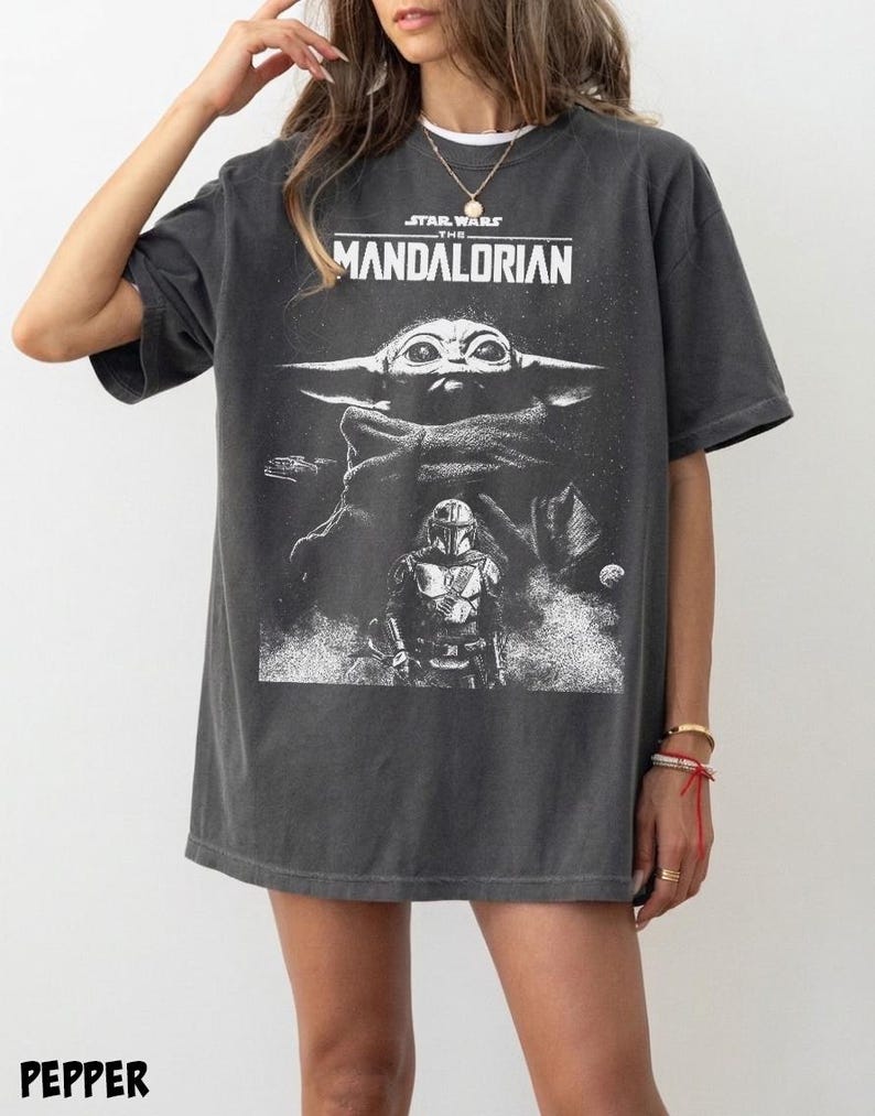 May include: Dark gray t-shirt featuring a graphic from the Star Wars series, "The Mandalorian." The design includes the show's title, a depiction of Grogu, and the Mandalorian. The word "PEPPER" is printed at the bottom of the shirt.