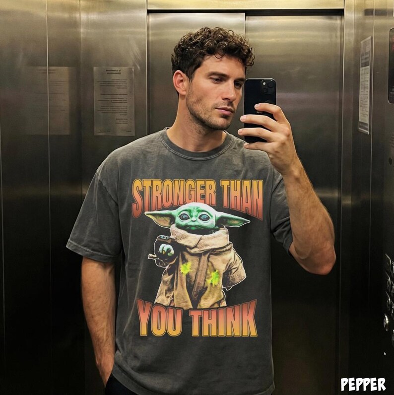 Pode incluir: T-shirt cinza escuro com um gr&aacute;fico de Grogu de The Mandalorian e o texto "STRONGER THAN YOU THINK" em laranja e amarelo. A camisa tem um visual vintage e desgastado.