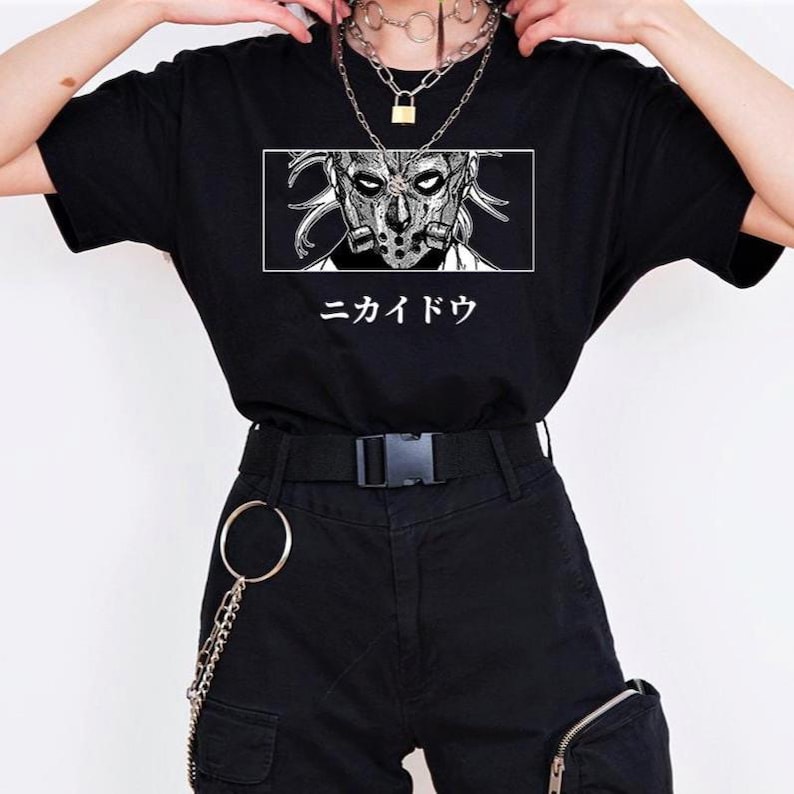 May include: Black t-shirt featuring a detailed black and white illustration of a masked character within a white rectangular frame. Japanese text is below the graphic. The shirt is paired with black shorts and accessories.