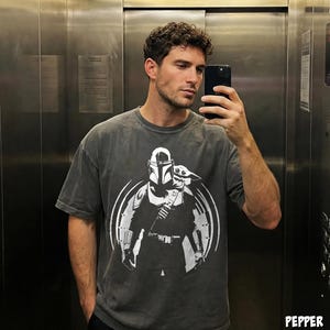May include: Dark grey t-shirt with a white graphic of The Mandalorian and Grogu. The design is circular, with the characters in the centre. The shirt has short sleeves and a crew neck. The shirt is made of a soft, comfortable material.